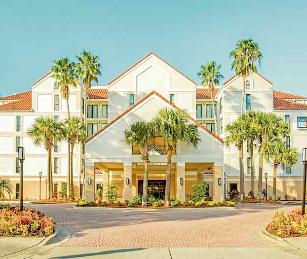 Kuva hotellista, jossa on valkoinen julkisivu, punaiset kattotiilet, ympäröity palmuilla ja hyvin hoidetuilla puutarhoilla. (Tekoälyn luoma teksti)