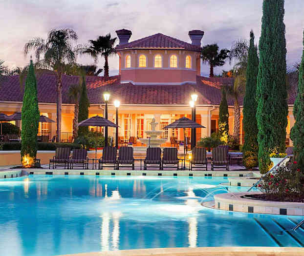 Ulkouima-allas, jonka ympärillä on aurinkotuoleja ja aurinkovarjoja valaistun huvilan edessä hämärässä. (Tekoälyn luoma teksti)