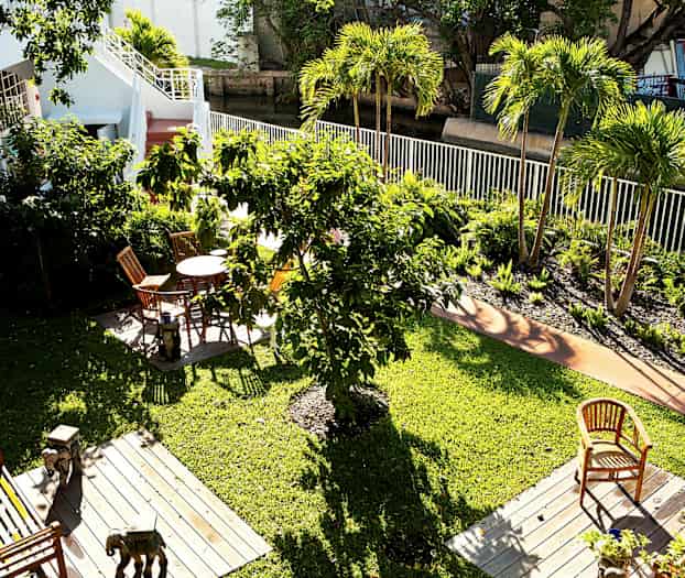 Näkymä vihreälle puutarhalle, jossa on useita puita ja kukkapenkkejä. Puutarhakalusteita on sijoitettu puisille alustoille nurmikolla. (Tekoälyn luoma teksti)