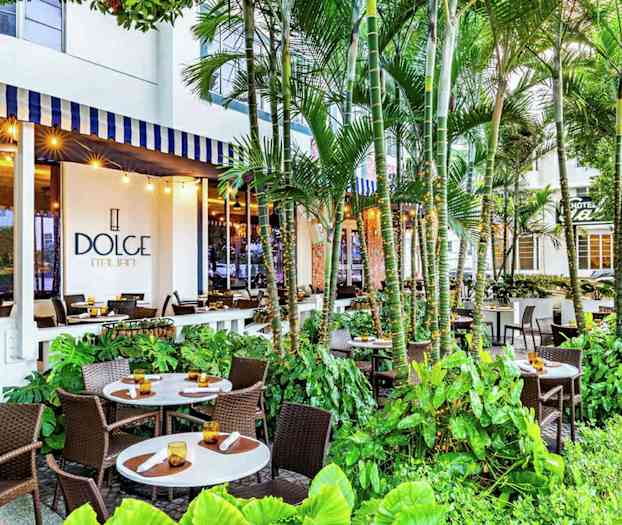 Ulkoterassi, jossa on useita pöytiä ja tuoleja rehevän kasvillisuuden keskellä, rakennuksessa näkyy kyltti 'Dolce Italian'. (Tekoälyn luoma teksti)