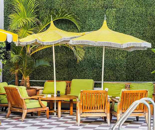 Ulkoistuimet, joissa vihreät tyynyt ja puupöytä, kelta-valkoraidallisten varjojen alla, taustalla kasvillisuutta. (Tekoälyn luoma teksti)