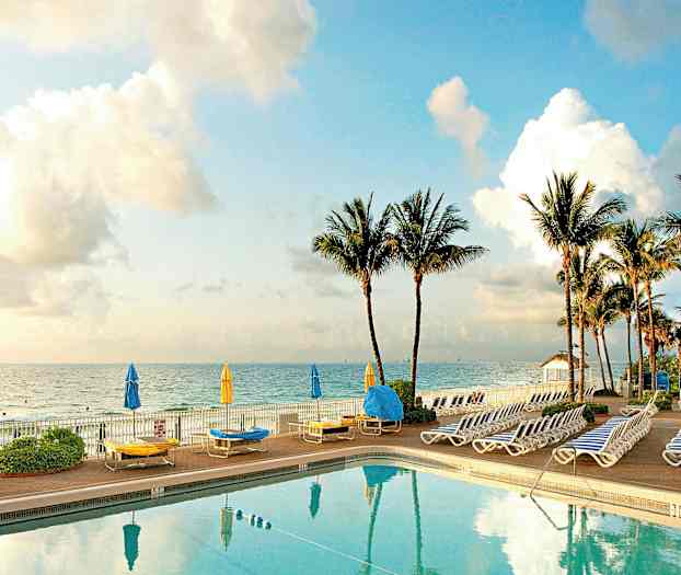 Uima-allas, jonka ympärillä on aurinkotuoleja ja aurinkovarjoja meren rannalla, taustalla palmuja. (Tekoälyn luoma teksti)