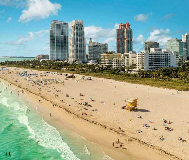 Hiekkaranta, jossa on ihmisiä uimassa ja auringossa meren äärellä. Taustalla näkyy korkeita rakennuksia. (Tekoälyn luoma teksti)