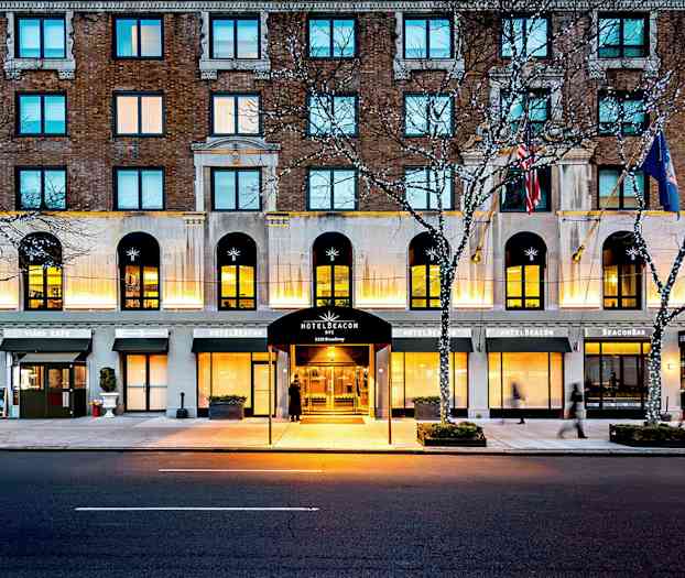 Hotellirakennus kadulla, sisäänkäynnillä, jossa lukee Hotel Beacon, ympärillä kahvilatunnelmaa ja valoja puissa. (Tekoälyn luoma teksti)