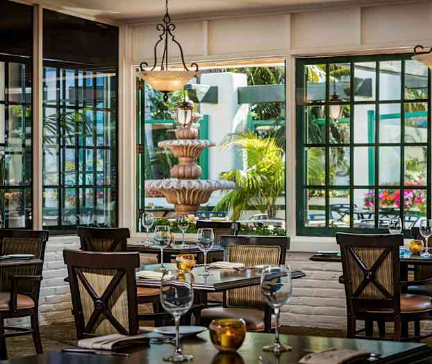 Kuva ravintolasalista, jossa pöydät ovat katettuina. Suuret ikkunat päästävät valoa sisään, ja ulkopuolella näkyy suihkulähde ja vihreitä kasveja. (Tekoälyn luoma teksti)