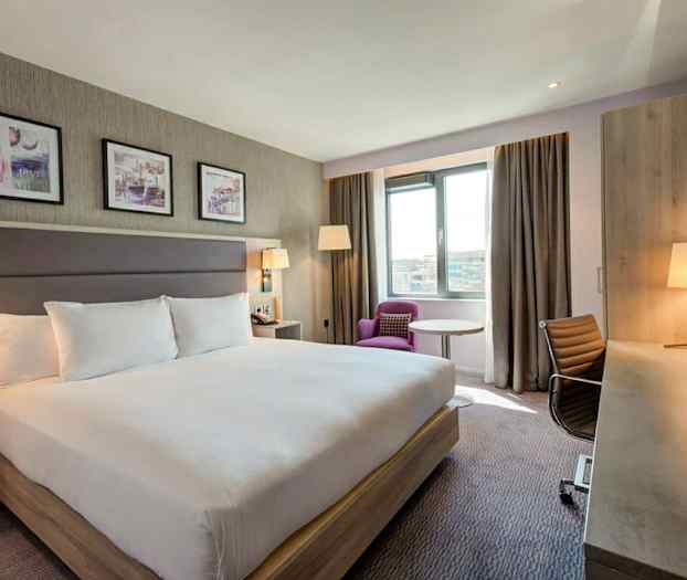 Hotellihuone, jossa on suuri parisänky, työpöytä, tuoli ja nojatuoli ikkunan vieressä. (Tekoälyn luoma teksti)