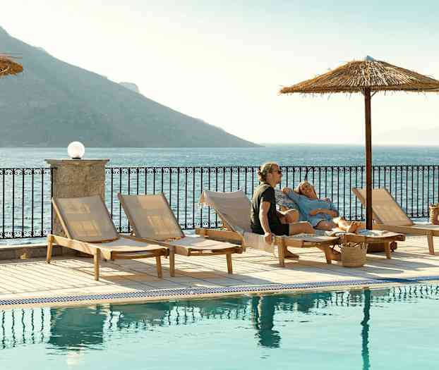 Aurinkotuoleja uima-altaan vieressä, näkymä merelle ja vuorille taustalla. (Tekoälyn luoma teksti)