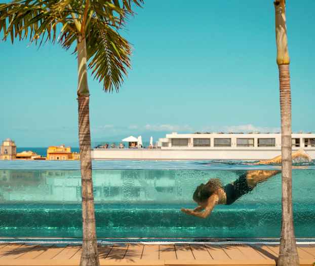 Henkilö ui veden alla läpinäkyvässä altaassa, taustalla rakennuksia ja palmupuu sinistä taivasta vasten. (Tekoälyn luoma teksti)