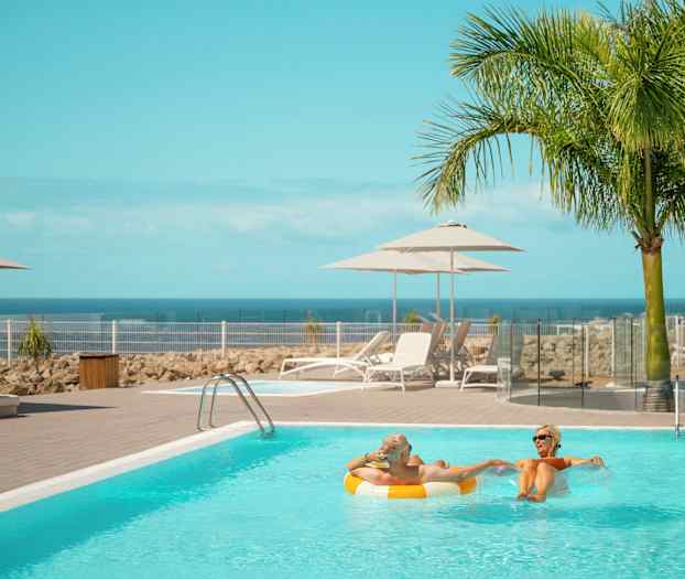 Uima-allas aurinkotuolien ja varjojen vieressä, taustalla meri ja palmut. (Tekoälyn luoma teksti)