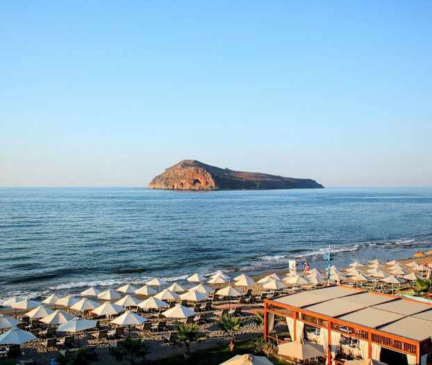 Ranta, jossa aurinkotuoleja ja varjoja, etualalla puurakenteinen kahvila ja taustalla näkyy kalliosaari. (Tekoälyn luoma teksti)