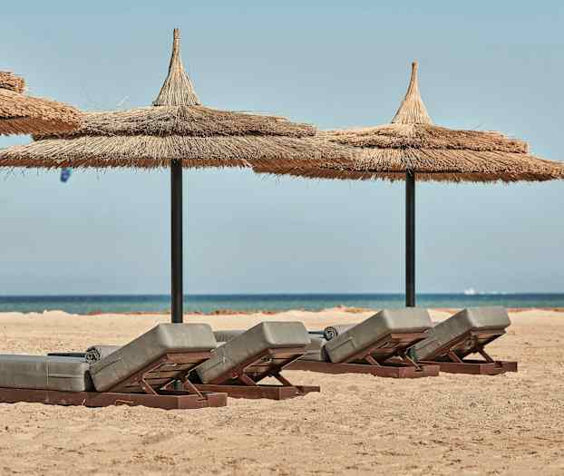 Aurinkotuolit olkikatolla hiekkarannalla, taustalla meri. (Tekoälyn luoma teksti)