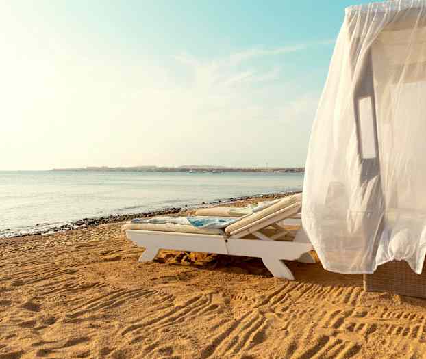 Aurinkotuoli, jossa valkoinen verho, hiekkarannalla meren äärellä. (Tekoälyn luoma teksti)