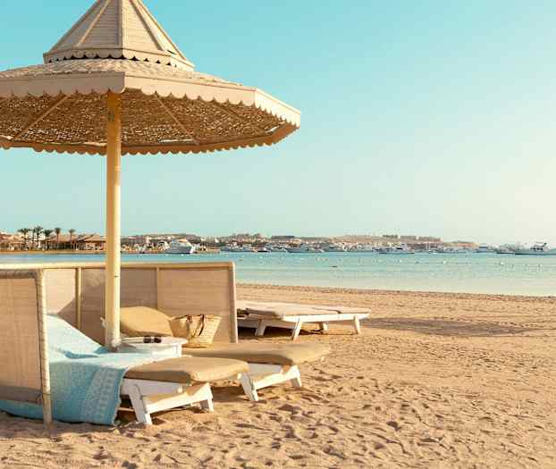 Ranta, jossa on aurinkotuoleja ja aurinkovarjoja sekä veneitä merellä. (Tekoälyn luoma teksti)