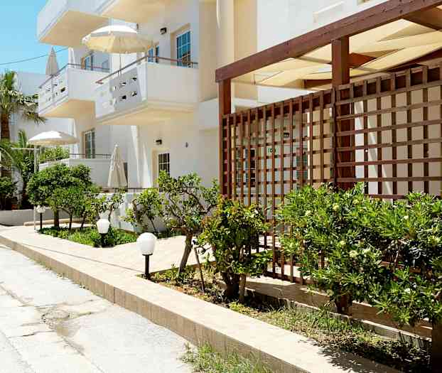 Katu, jolla on valkoisia rakennuksia, vihreitä pensaita ja aurinkovarjoja. (Tekoälyn luoma teksti)