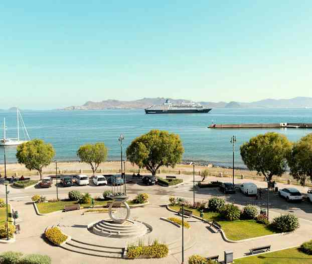 Näkymä satamaan, jossa etualalla parkkipaikka ja puita, veneitä ja risteilyalus sinisellä merellä, sekä vuoret taustalla. (Tekoälyn luoma teksti)