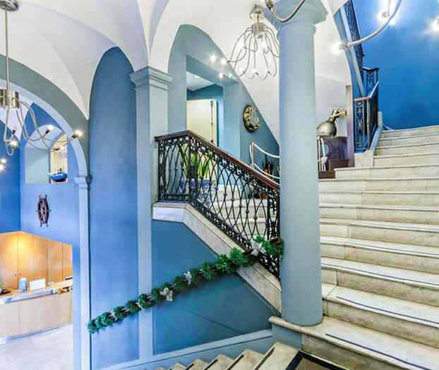 Sisätilakuva aulasta, jossa on vaaleasta marmorista valmistetut portaat ja siniset seinät, koristeltuna vihreillä köynnöksillä. Portaissa on kaiteet mustasta metallista ja puusta. (Tekoälyn luoma teksti)