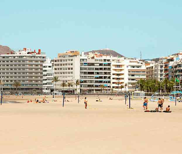 Ranta, jossa hiekkaa ja ihmisiä, jotka ottavat aurinkoa ja pelaavat lentopalloa useiden kerrostalojen edessä. (Tekoälyn luoma teksti)