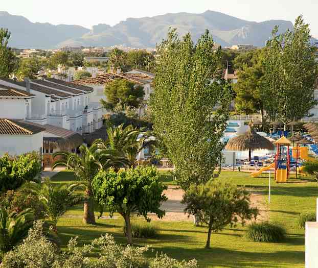 Näkymä viheralueelle, jossa on puita, nurmikkoa ja pieniä valkoisia rakennuksia sekä taustalla vuoria. (Tekoälyn luoma teksti)