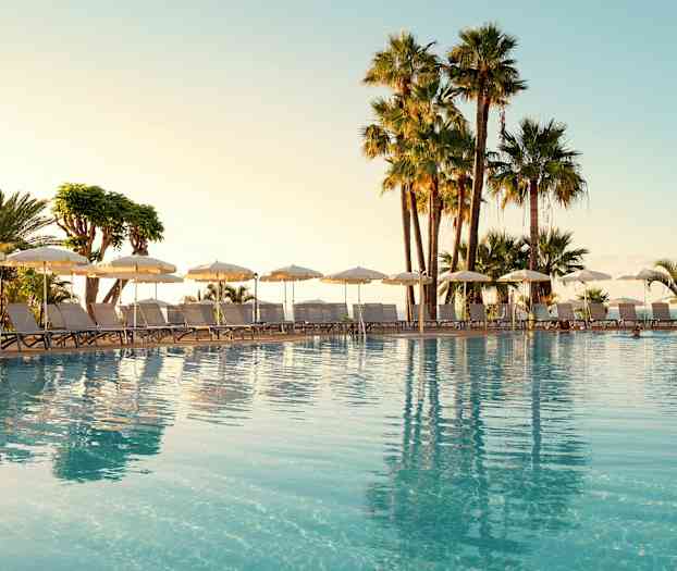 Ulkoallas, aurinkotuoleja ja aurinkovarjoja, ympärillä palmuja. (Tekoälyn luoma teksti)