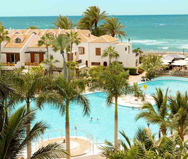 Hotellirakennukset terrakotanvärisillä katoilla uima-altaan vieressä, ympäröitynä palmuilla ja merellä taustalla. (Tekoälyn luoma teksti)