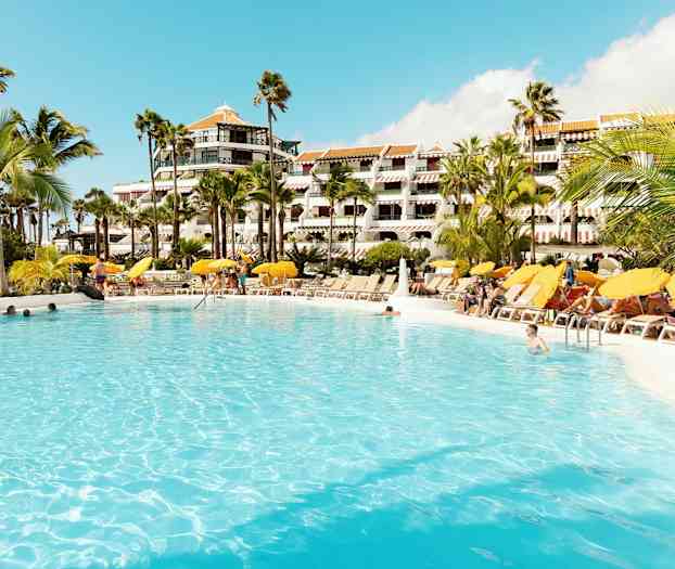 Suuri uima-allas, jonka ympärillä aurinkotuoleja ja aurinkovarjoja hotellin edessä. (Tekoälyn luoma teksti)