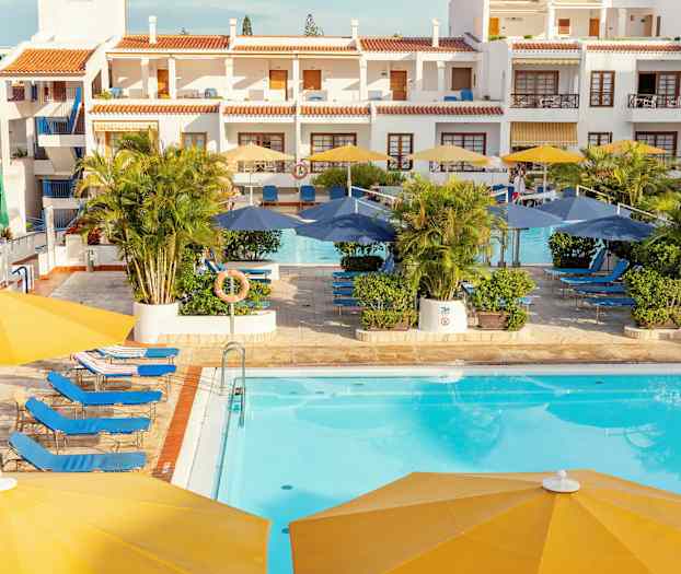 Hotellin allasalue, jossa on aurinkotuoleja, aurinkovarjoja ja rakennuksia taustalla. (Tekoälyn luoma teksti)