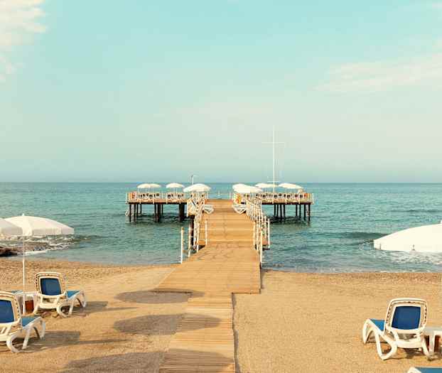 Hiekkaranta aurinkotuoleilla ja varjoilla, laituri joka johtaa vesialueelle. (Tekoälyn luoma teksti)