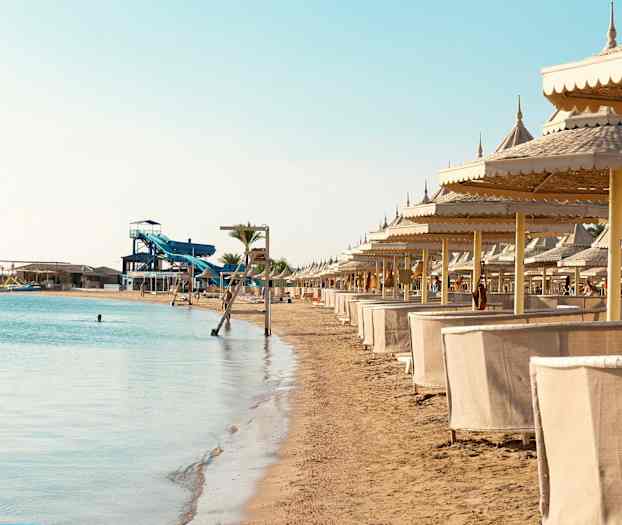 Hiekkaranta, jolla on rivi aurinkovarjoja ja cabana-majoja meren rannalla, taustalla vesiliukumäki. (Tekoälyn luoma teksti)