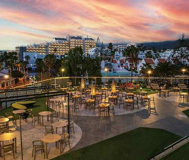 Ulkoterassi, jossa pöytiä ja tuoleja iltavalaistuksessa. Taustalla rakennuksia ja värikäs auringonlasku. (Tekoälyn luoma teksti)