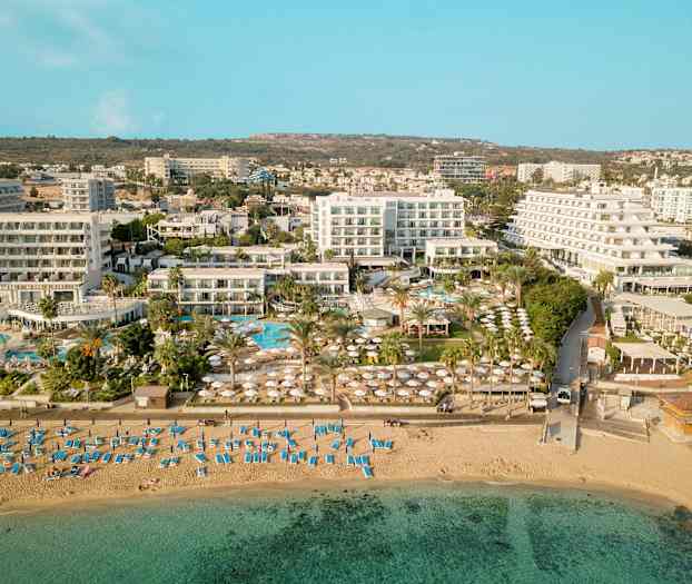 Ilmakuva, joka näyttää rannan aurinkotuoleilla ja aurinkovarjoilla sekä hotellirakennukset ja allasalueen taustalla. (Tekoälyn luoma teksti)