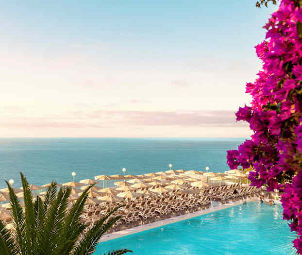 Aurinkotuoleja ja aurinkovarjoja uima-altaan äärellä, merinäkymä ja kukkia. (Tekoälyn luoma teksti)