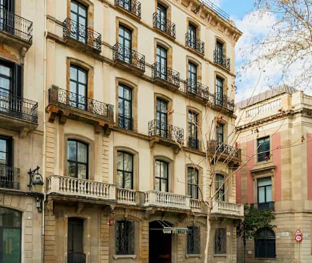 Rakennuksen julkisivu, jossa on useita kerroksia ja takorautaparvekkeita, sijaitsee kadun varrella puiden ja jalkakäytävän vieressä. (Tekoälyn luoma teksti)