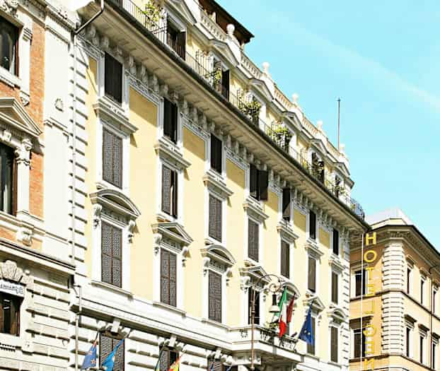 Monikerroksisen hotellin julkisivu, koristeltu lipuilla ja ympäröity autoilla ja moottoripyörillä. (Tekoälyn luoma teksti)