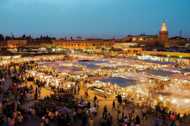 Jemaa el Fna -tori Marrakechin tunnelmallisessa iltahämärässä. Valaistujen ruokakojujen ympärillä on paljon ihmisiä.