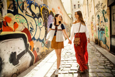 Kaksi nuorta naista kävelee käsi kädessä graffitien peitossa olevien seinien reunustamaa nupukivikatua.