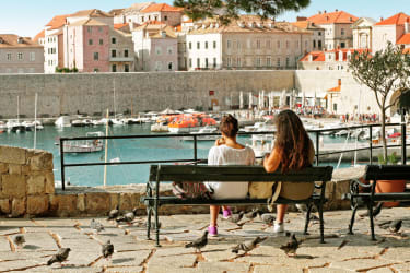 Kaksi naista istuu puistonpenkillä ja katsoo edessä kohoavaa Dubrovnikin Vanhaakaupunkia.