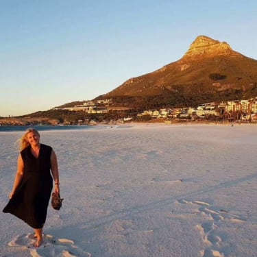 Jutun kirjoittaja kävelee iltavalaistuksessa hiekkarannalla mustassa kesämekossa, sandaalit kädessä.