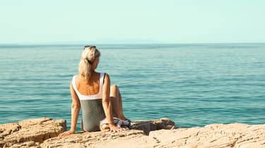 Nainen uimapuvussa istuu rantakivillä ja katsoo merelle.