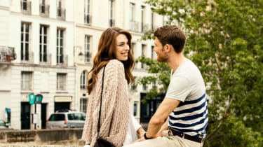 Mies ja nainen katsovat toisiaan hymyillen, taustalla vehreä puu ja vaalea kaupunkitalo takorautaparvekkeineen.