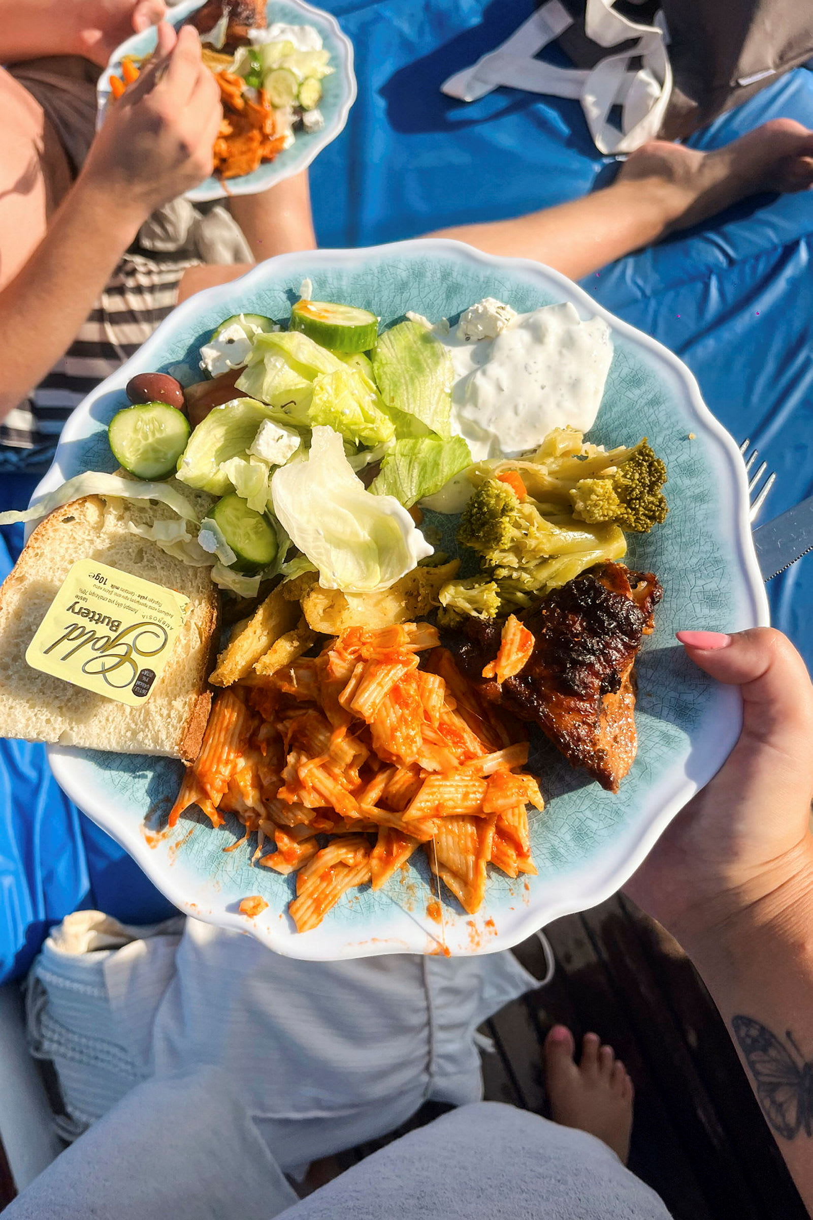 Lautasella ruokaa, jossa on tomaattikastiketta sisältävää pastaa, vihanneksia ja fetajuustoa sisältävää salaattia, parsakaalia, lihapala ja pala leipää voin kanssa. Taustalla näkyy toinen lautanen sekä henkilö sinisellä pöytäliinalla. (Tekoälyn luoma teksti)