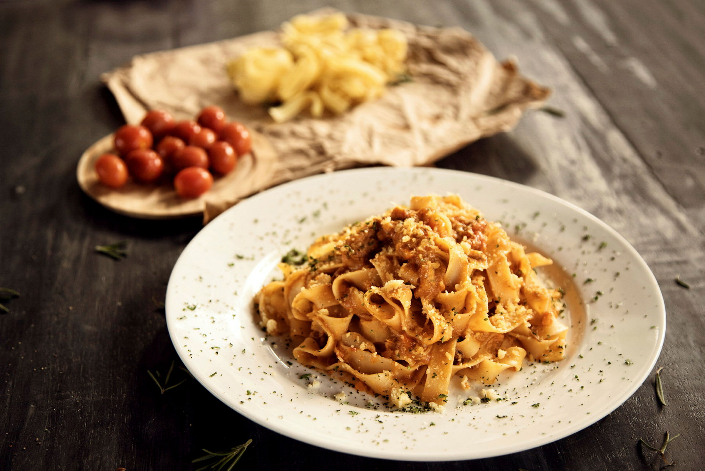 Lautanen pastaa kastikkeella ja yrteillä, taustalla kirsikkatomaatteja ja raakaa pastaa. (Tekoälyn luoma teksti)