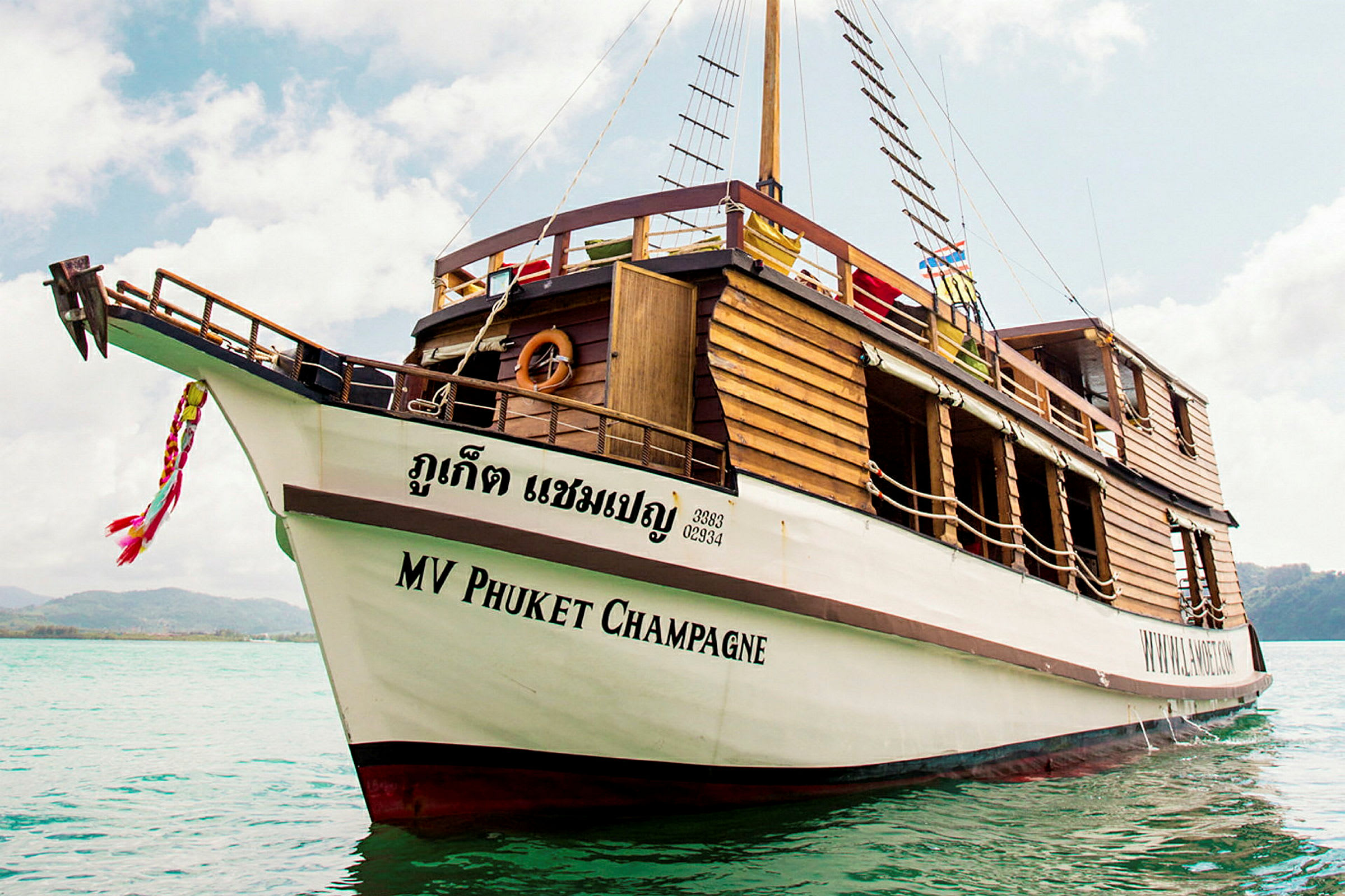 Puusta tehty vene, jonka kyljessä lukee 'MV Phuket Champagne', ankkuroituna kirkkaaseen veteen. (Tekoälyn luoma teksti)
