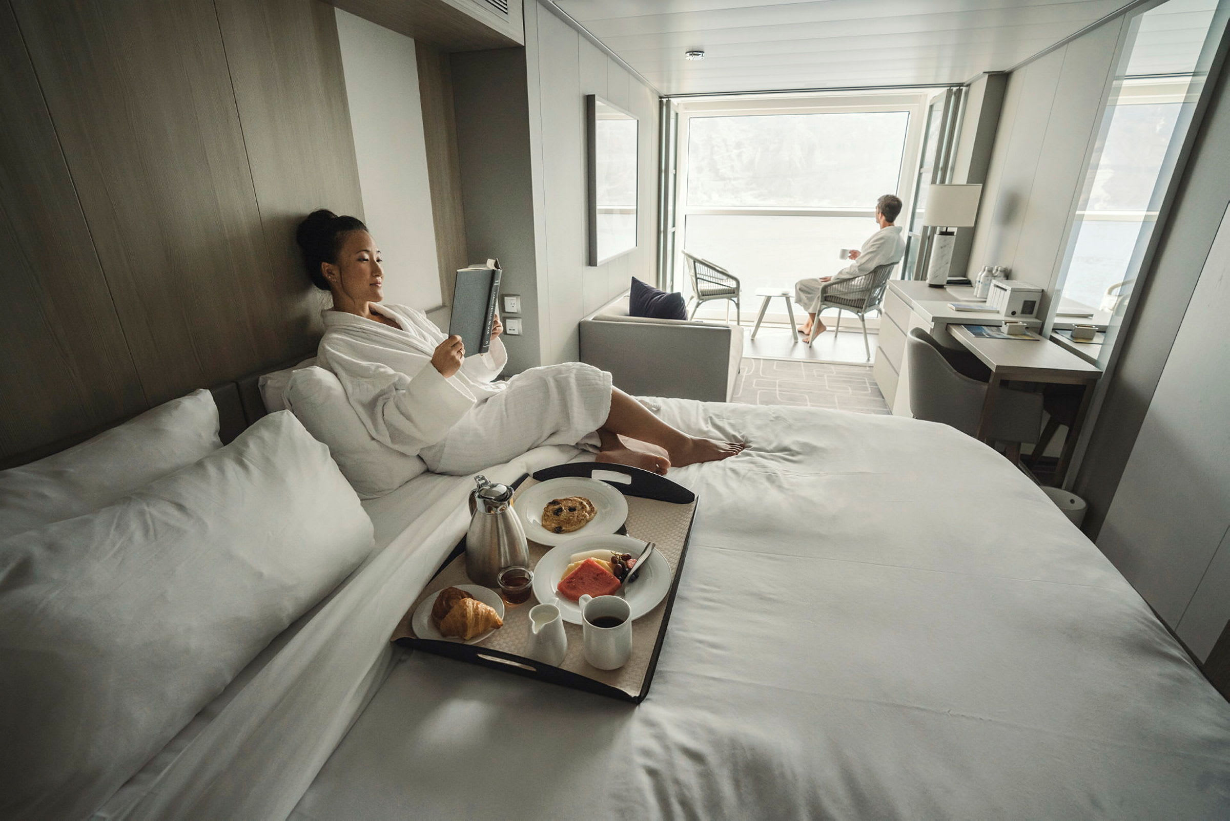 Henkilöitä rentoutuu valoisassa ja modernissa hotellihuoneessa, jossa aamiainen on tarjoiltu sängyllä ja istumapaikka suuren ikkunanäkymän äärellä. (Tekoälyn luoma teksti)