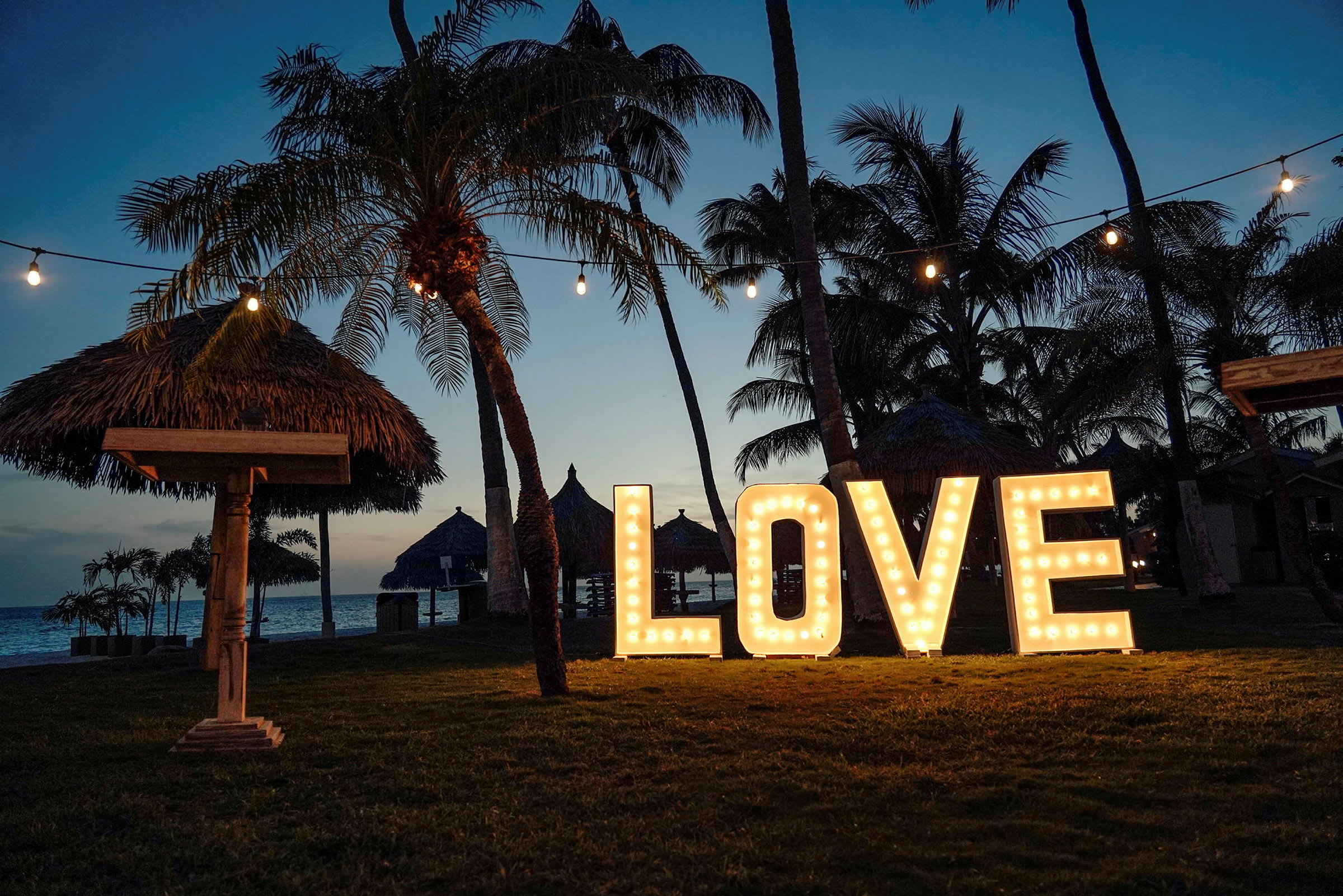 Suuret valaisevat kirjaimet, jotka muodostavat sanan 'LOVE', ovat nurmikolla palmujen ympäröimänä. Luonnonmaiseman yläpuolella roikkuvat valoketjut, ja taustalla näkyy ranta hämärässä. (Tekoälyn luoma teksti)