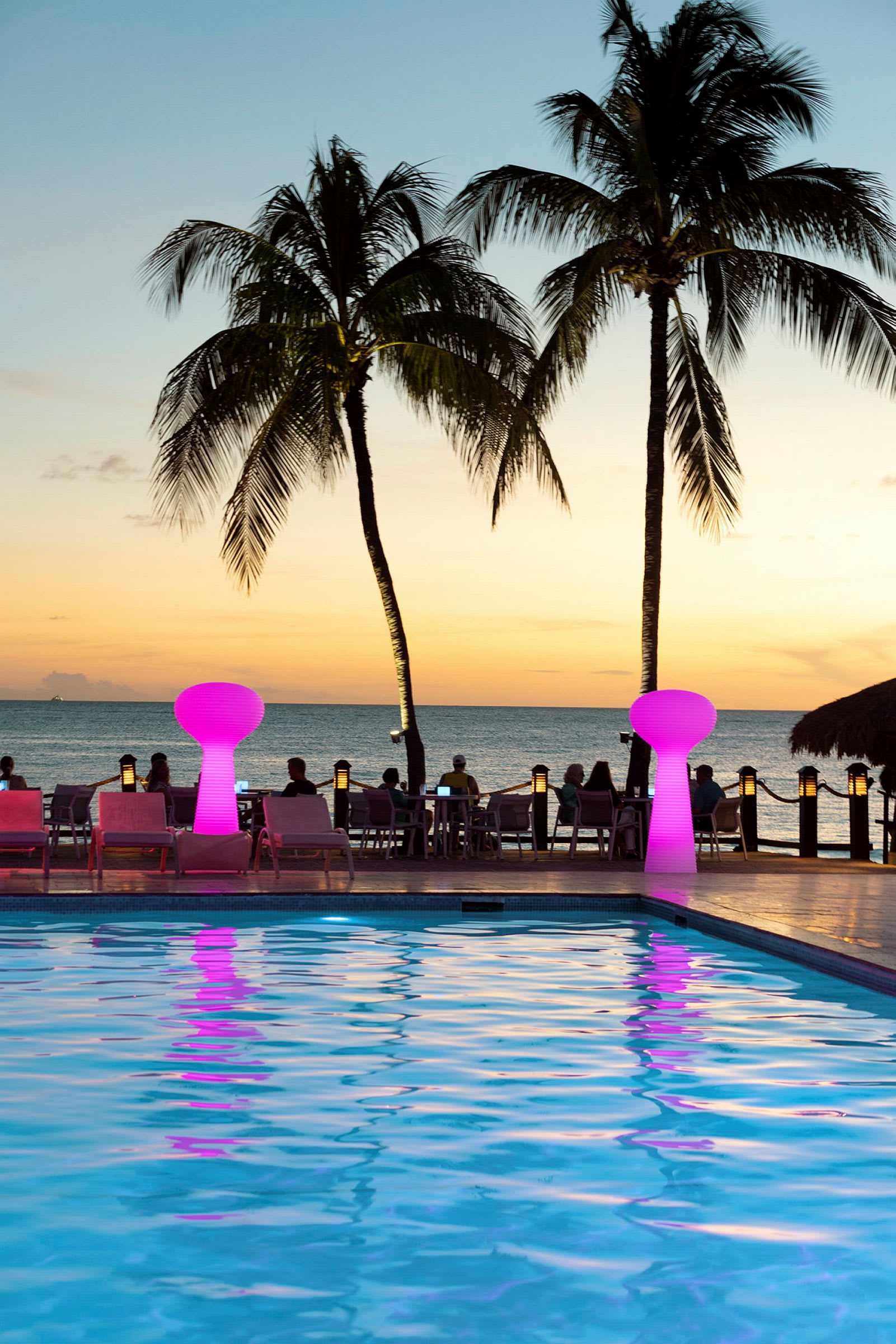 Auringonlasku meren äärellä palmujen, altaan ja värikkäiden lamppujen kanssa. (Tekoälyn luoma teksti)