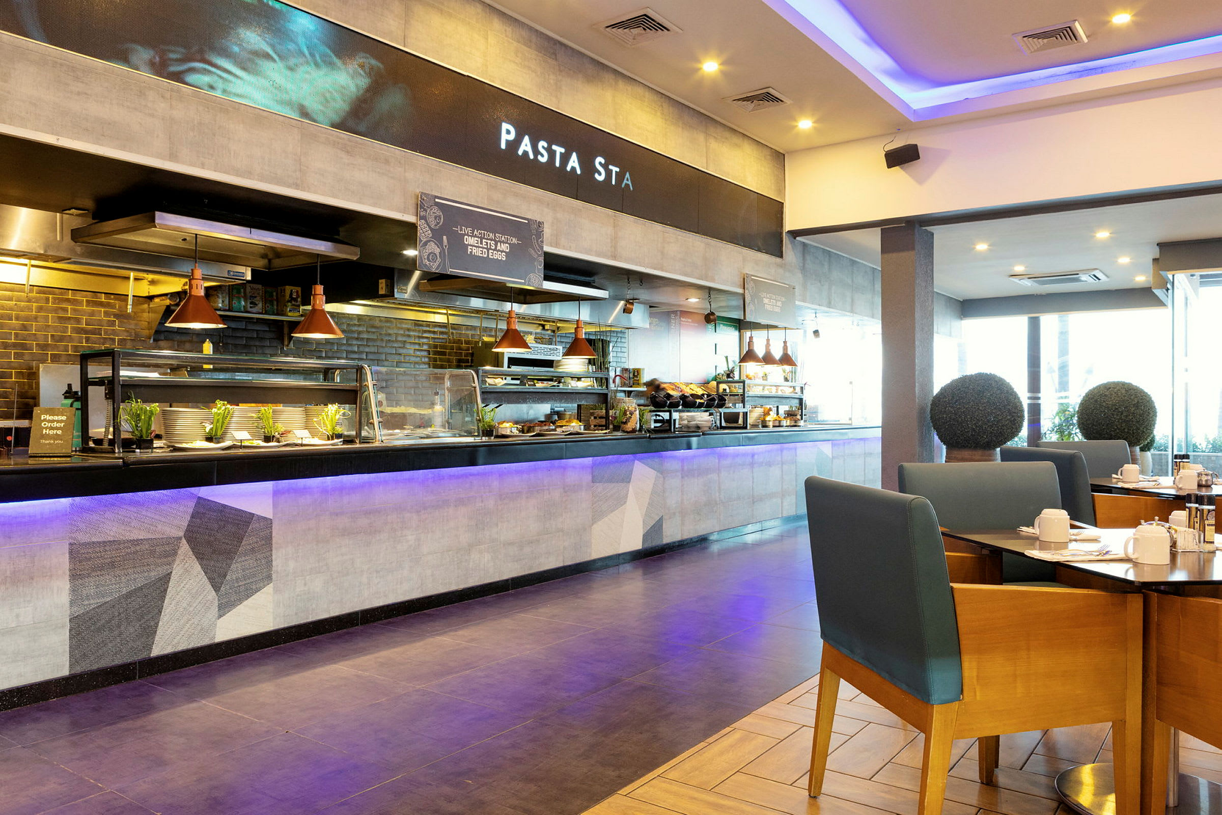 Ravintola, jossa on avoin tarjoilutiski ja istumapaikkoja. Etualalla näkyy pöytiä ja tuoleja, ja tarjoilutiskin takana on kyltti, jossa lukee 'Pasta Sta'. (Tekoälyn luoma teksti)