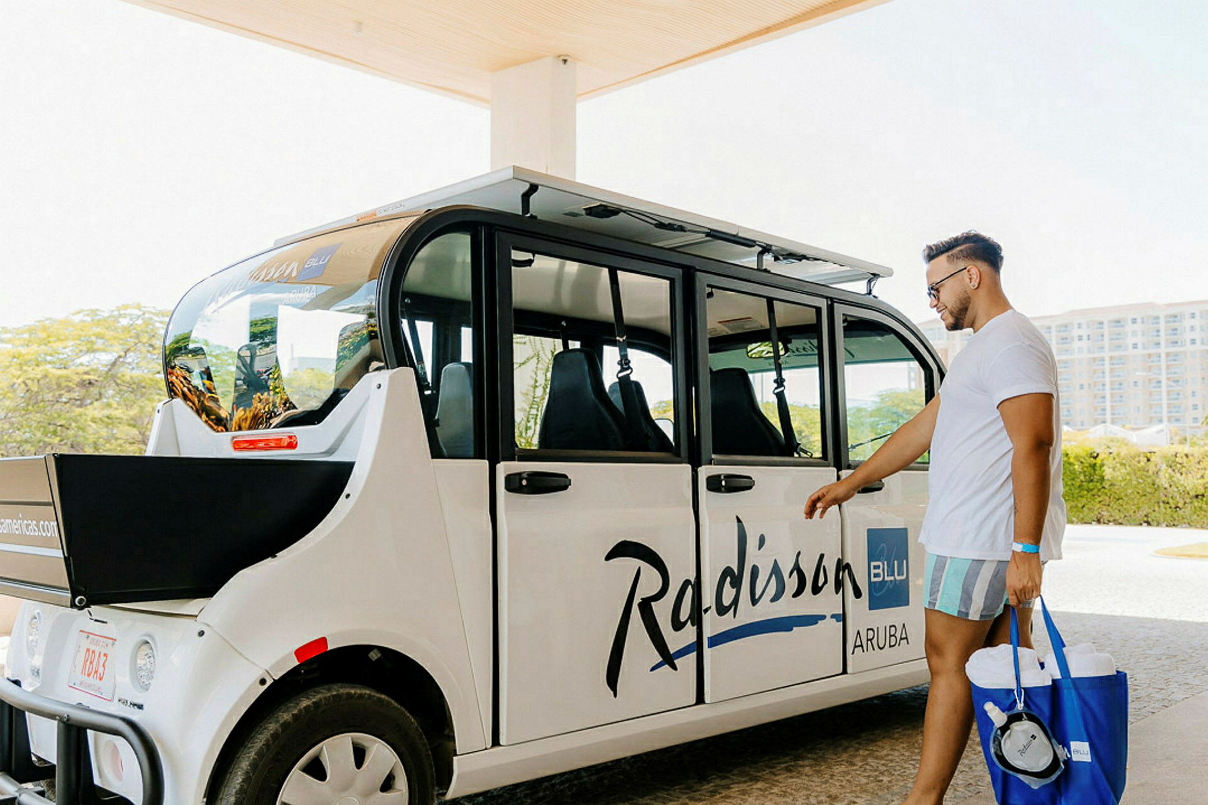 Turisti seisoo valkoisen kuljetusauton vieressä, jossa lukee 'Radisson Blu Aruba'. (Tekoälyn luoma teksti)