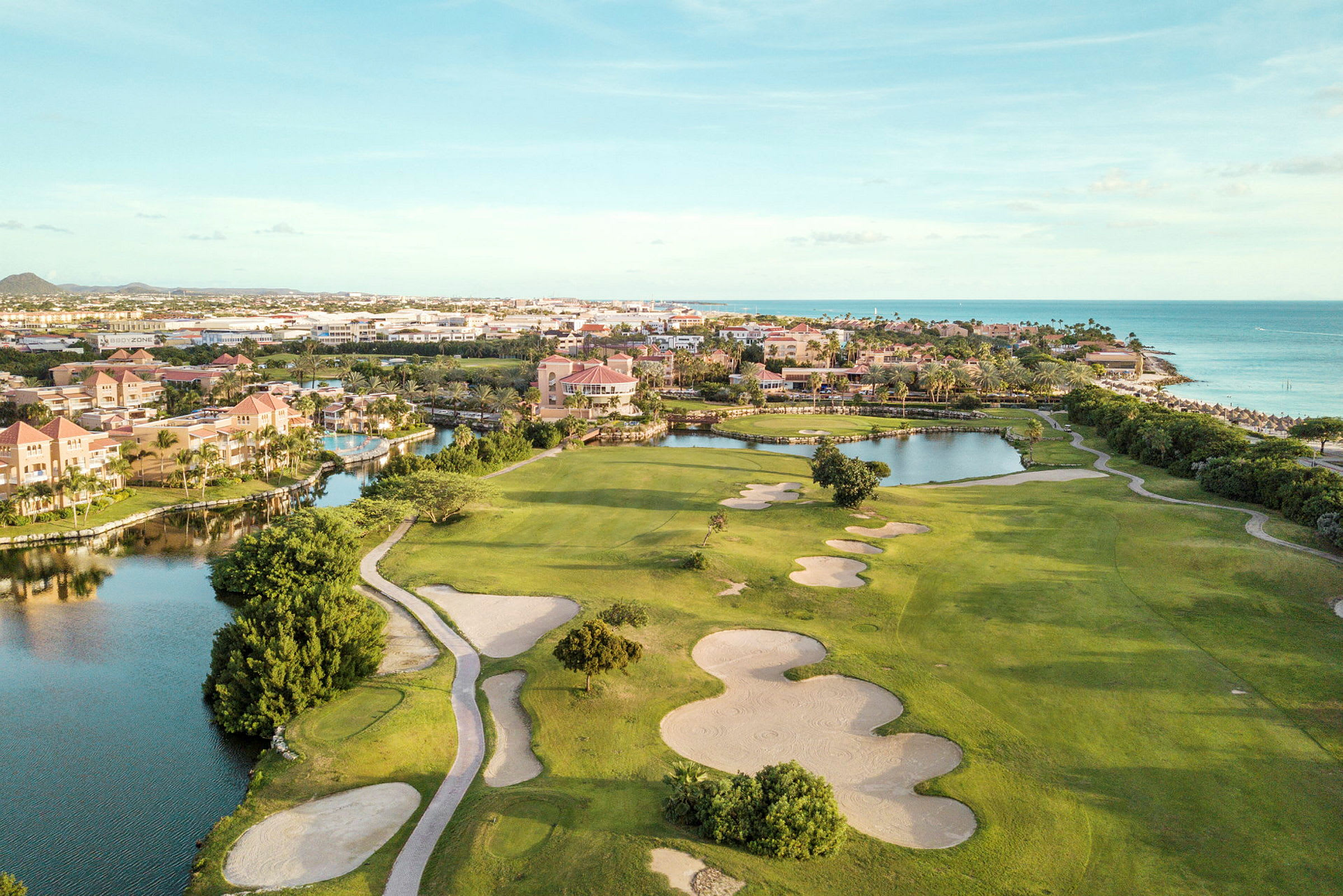 Golfkenttä, jossa on hiekkabunkkereita ja vihreitä nurmialueita lähellä vesiväylää ja rantaa. Taustalla rakennuksia. (Tekoälyn luoma teksti)