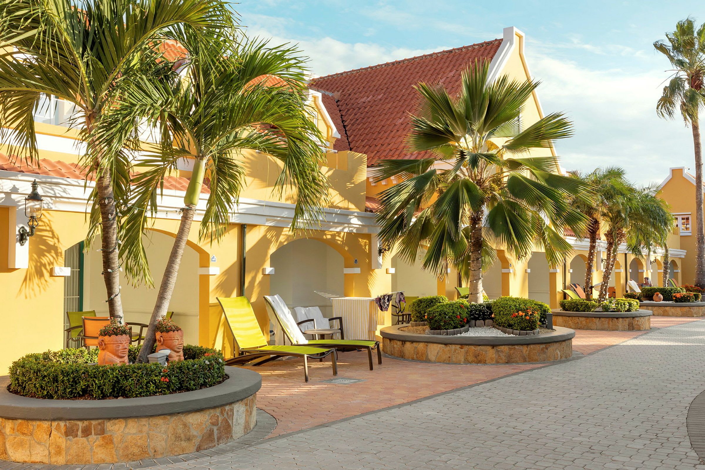 Keltainen rakennus punaisilla katoilla, ympäröitynä palmuilla ja kivetyllä kävelytellä. (Tekoälyn luoma teksti)