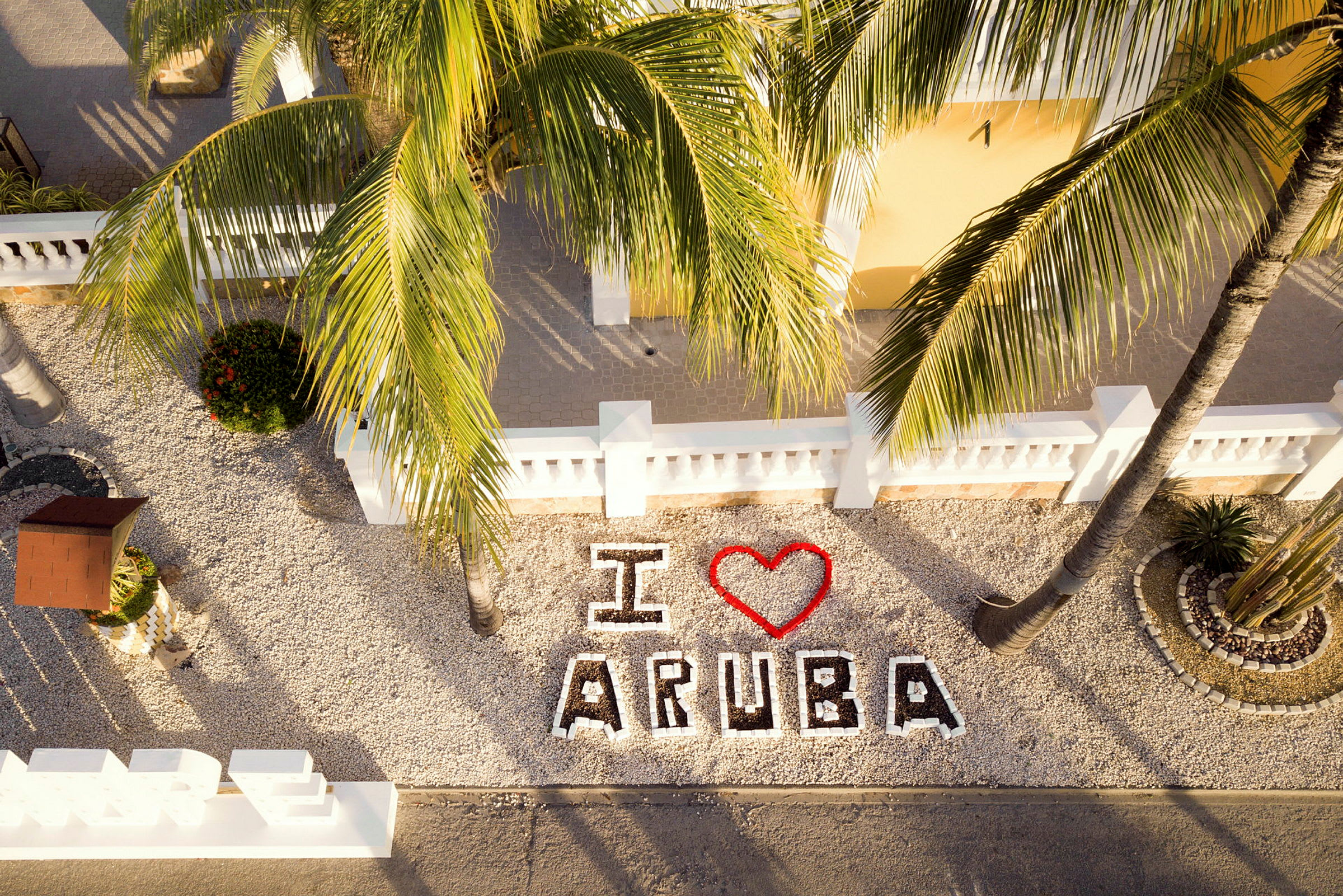 Teksti "I ❤️ Aruba" näkyy koristeellisten kiviasetelmien ja palmujen joukossa. (Tekoälyn luoma teksti)